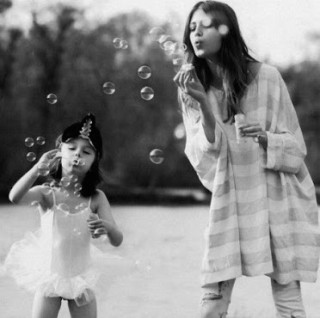 mãe com criança de uns 4 anos brincando em um parque de fazer bolhas de sabão. A menina veste uma roupa de ballet, com saia rodada e meia calça