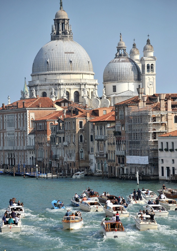 Vista da cidade italiana de Veneza, no dia do casamento do ator George Clooney, nesta imagem , com céu lindo, azul claro, ao fundo a cidade com prédios antigos, destacando-se as cúpulas enormes cinzas, todas as ruas são canais e inúmeros barcos lotados de jornalistas navegam acompanhamento o barco dos noivos.