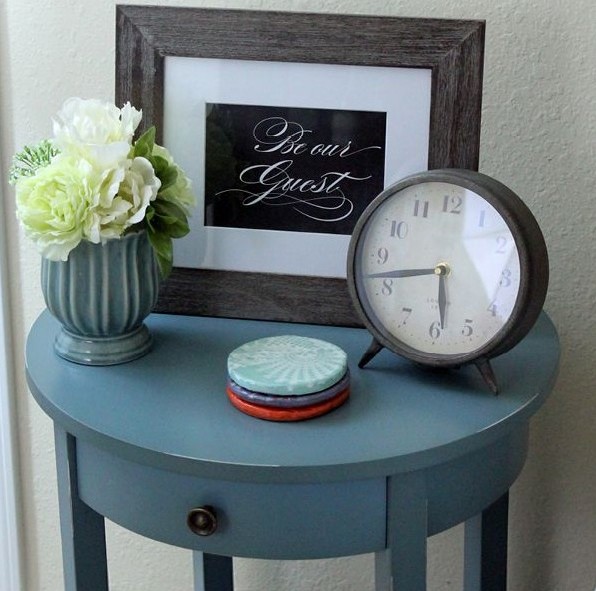 criado mudo azul. Em cima uma vasinho tb azul com flores frescas brancas, um relógio de ponteiros e um quadrinhos escrito be our guest