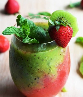Copo baixo com sucos misturados: de goiaba vermelho e de kiwi verde formando efeito bicolor. Sobre a borda suco folhas de hortelã e um morango enfeitam e dão um toque de mais cor ao drinque