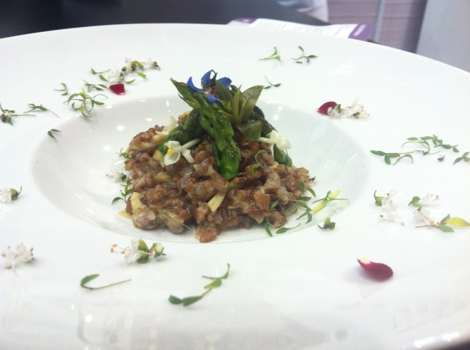 em um prato grande de porcelana branca está colocado no centro um punhado de risoto de arroz integral com pontas de aspargo verde enfeitando em cima. Na borda de aba larga do prato pequenas folhas de ervas verdes enfeitam o prato com pétalas vermelhas de mini rosas formando um contraste delicado.