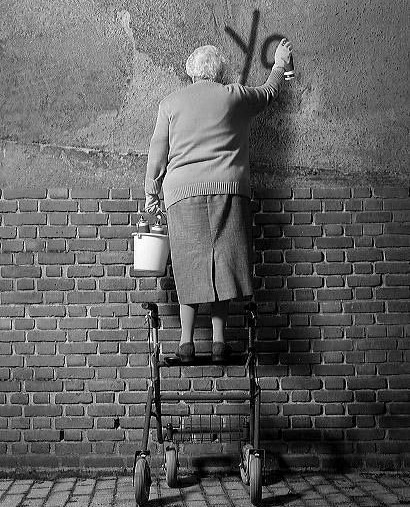 idosa com cabelinhos curtos e brancos em cima de uma escada pichando um muro