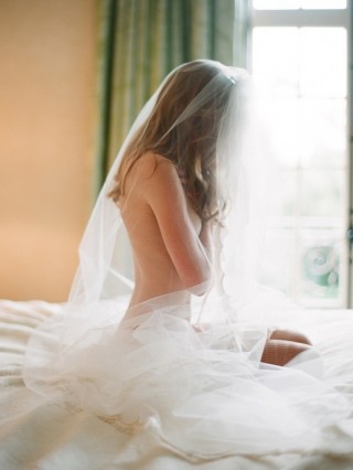 Moça sem roupa apenas com o véu de noiva sentada nua sobre a cama. O rosto está escondido e pela janela do quarto entra uma luminosidade que infere que está no início da manhã. O cenário é de intimidade e paz.