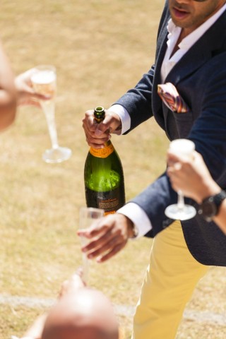 Em ambiente aberto, um homem vestindo camisa branca e blaser azul, oferece champanhe Veuve Clicquot, onde aparece mãos de pessoas segurança a taça "flute" com champanhe. 