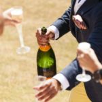 Em ambiente aberto, um homem vestindo camisa branca e blaser azul, oferece champanhe Veuve Clicquot, onde aparece mãos de pessoas segurança a taça "flute" com champanhe.