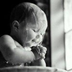 Foto em preto e branco, de uma bebê com 6 meses, recebendo o batismo - a água escorre pela cabeça e ombros nus , enquanto ele aperta as mãos juntas como se estivesse em prece.