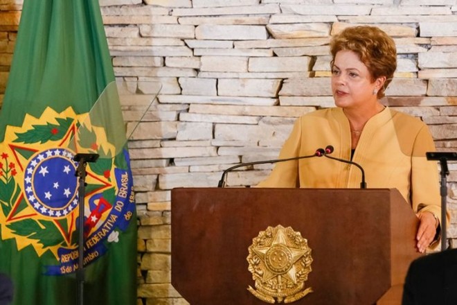 A Presidente Dilma Rousseff está atrás do púlpito presidencial flanco a frente de dois microfones . Atrás dela, a esquerda está a bandeira da República e logo a frente percebe-se um pedestal de girafa com a placa de vidro do teleprompter de onde ela está lendo a fala.