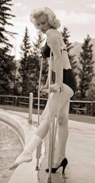 Marilyn Monroe, está em pé na borda de uma piscina, ela está usando um proteção no tornozelo direito e usa uma muleta para se sustentar. Ela veste um biquini preto.