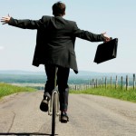 homem de costas, de terno, andando numa bicicleta numa estrada, ela está com os braços aberto, e na mão direita segura uma mala de executivo. Ao fundo um céu azul claro num dia de sol.