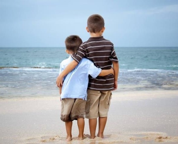 a foto mostra dosi meninos, de 7 e 5 anos abraçados olhando o mar. Eles estão com os pés na areia. a imagem transmite muito carinho entre os doi