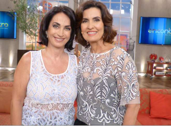 Claudia Matarazzo ao lado de Fátima Bernardes no Programa Encontro da Rede Globo, Claudia está a esquerda usando blusa rendada, na cor branca com detalhes em azul, e Fátima uma blusa transparente com detalhes bordados e um top preto por baixo. 