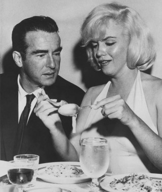 Atriz Marilyn Monroe, numa mesa de jantar, tenta comer macarrão usando garfo e colher, ao seu lado um homem de terno escuro está olhando para o gesto dela. Na mesa além dos pratos , tem uma taça com água e outra com vinho tinto. Ela usa um vestido claro, preso no pescoço e sem mangas.