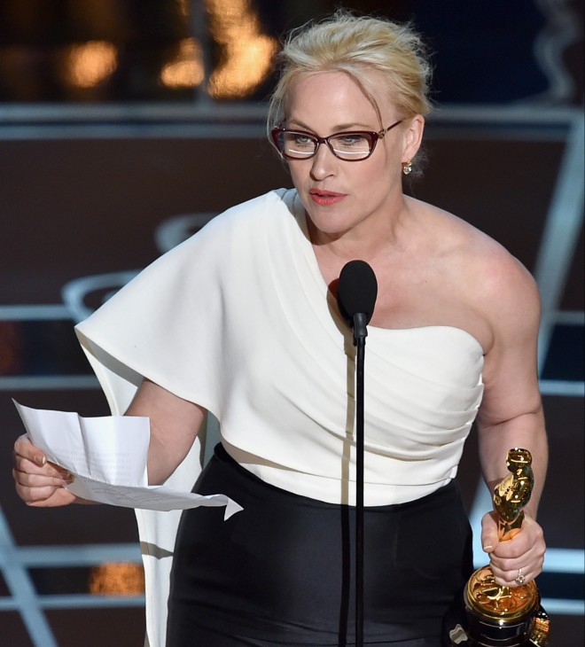 Atriz Patrícia Arquette, durante a Premiação do Oscar 2015, veste saia longa na cor preta, uma blusa branca, com o ombro esquerdo a mostra, está diante do microfone fazendo seu discurso de agradecimento. Na mão direita segura um papel e na outra mão segura a estatueta do Oscar.