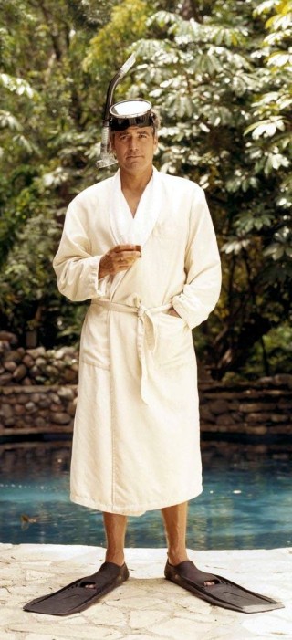 O ator George Clooney, junto a uma piscina, veste um roupão , na cor branca, usa óculos de mergulho na testa e um pé de pato, para mergulho.
