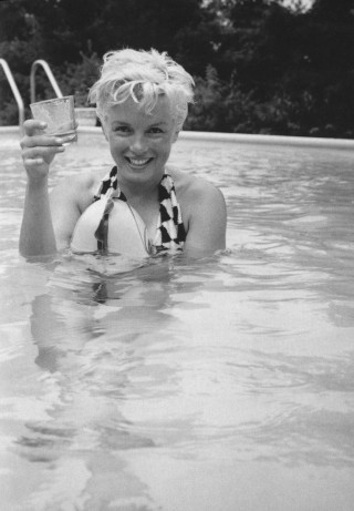 Imagem em preto e branco, Atriz Marilyn Monroe, com um maiô, está dentro de uma piscina, mas na sua mão direita tem um copo de vidro.