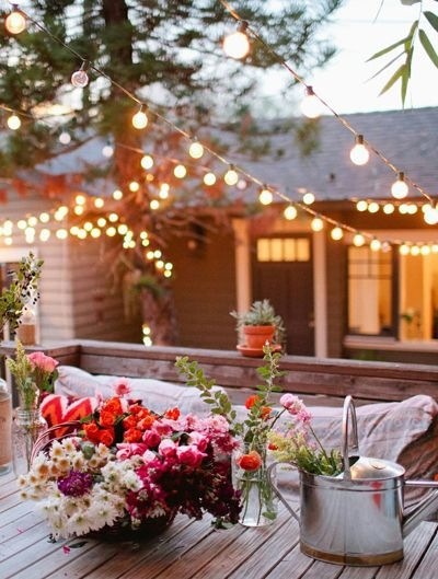 Imagem de casa no campo, num dia lindo, em frene a ela um ciprestre. No jardim uma mesa florida, com um regador servindo de vaso e com flores. No ar muitos fios com lâmpadas acesas.