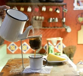 uma xícara de cafezinho de ágata branca está em primeiro plano com um bule também em ágata branco vertendo café fumegante em um coador de pano individual por onde o café passa e cai na xícara. Ao fundo percebe-se um ambiente de cozinha rústico e acolhedor com paredes pintadas de laranja .