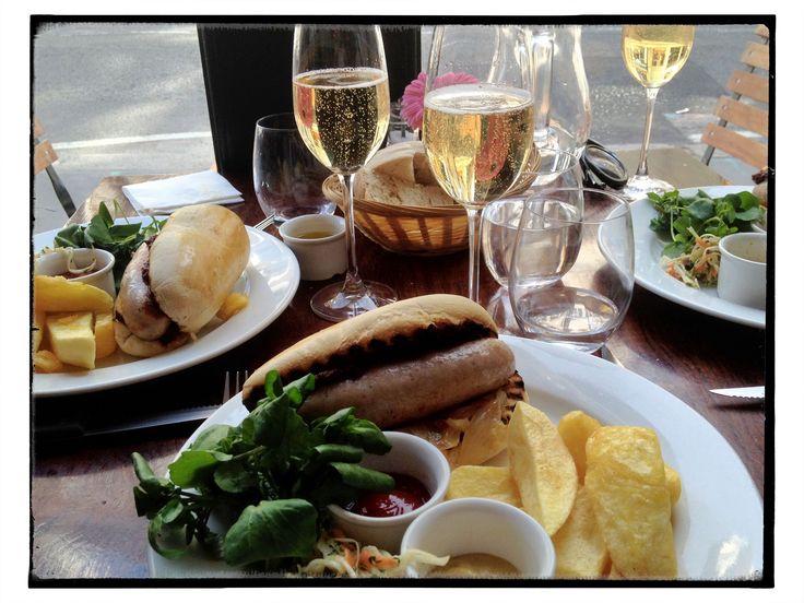 Sobre uma mesa em madeira lista estão dois pratos com um apetitoso cachorro quente com a salsicha devidamente grelhada com aparência chocante aparecendo dentro do pão semi tostado. No prato, complementam o hot dog, batatas fritas e duas mini cumbucas brancas com ketchup e mostarda, além de uma salada de agrião. Além dos copos de água, ha também taças de champanhe borbulhante e dourado convidando a um brinde.