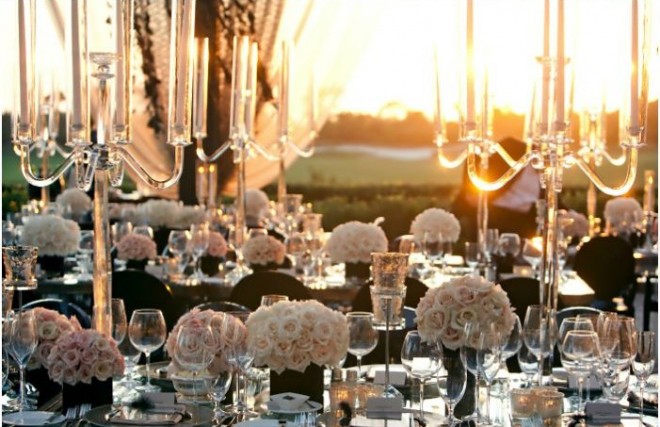 Uma mesa com candelabros de cinco braços em prata e muitos vasos baixos de rosas brancas está colocada a frente de uma janela. Através dela se vê a luz de por do sol que acentua a beleza da pradaria de das taças de cristal colocadas nos lugares dessa mesa formal de casamento. A imagem é de requinte e aconchego enfatizada pelo luz quente de final de tarde.