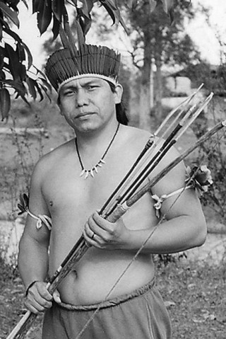 indio Daniel Munduruku, na mata segurando arco e flechas, A foto é preto e branca, na cabeça ela usa o cocar indígena, um colar com dentes de animais e nos braços usa junto ao bícce