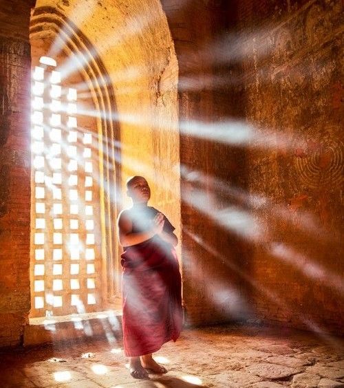 monge budista tibetano, vestindo seus mantos sagrados na cor marsala, está de pé com as mãos unidas junto ao peito junto a uma ampla janela do tempo por onde entra uma grande luz.