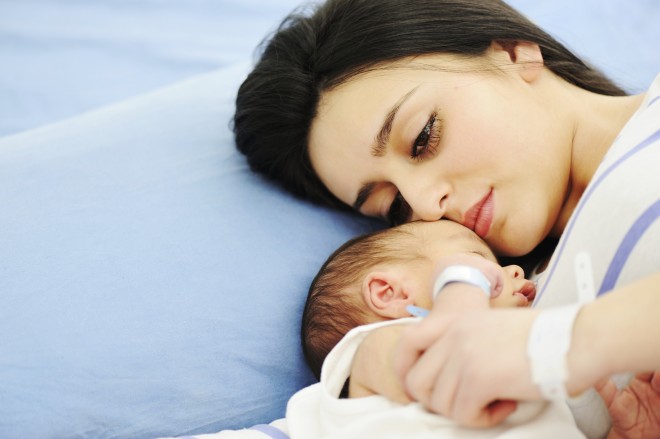 mãe jovem, deitada numa cama com lençóis azuis , abraçada ao seu bebê que dorme profundamente. 
