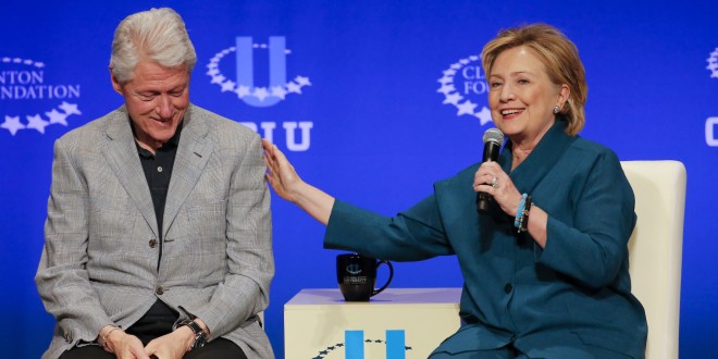 Bill Clinton ao lado de Hillary Clinton, durante uma entrevista na Fundação Bill Clinton, ambos estão sentados, ela está a esquerda dele. Ele veste uma camisa azul escura e blazer cinza claro. Ela está vestindo um conjunto azul petróleo e está com o microfone na mão esquerda.