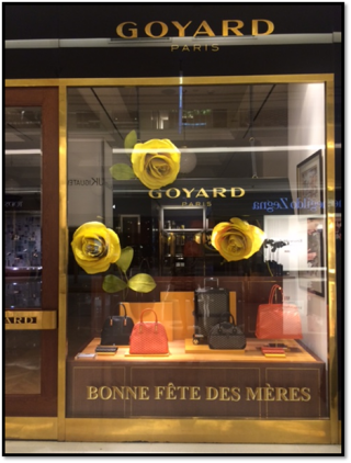 vitrine de uma loja chamada Goyard Paris, onde bolsas em tons coral e preta, estão junto a três rosas enormes amarelas. Embaixo da vitrine está a inscrição: Bonne Fête des mères em letras douradas.