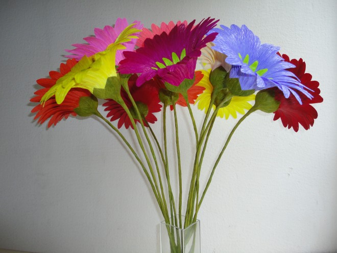 num vaso de cubo de vidro transparente, estão flores coloridas, em tons azul, vermelho, amarelo, rosa.