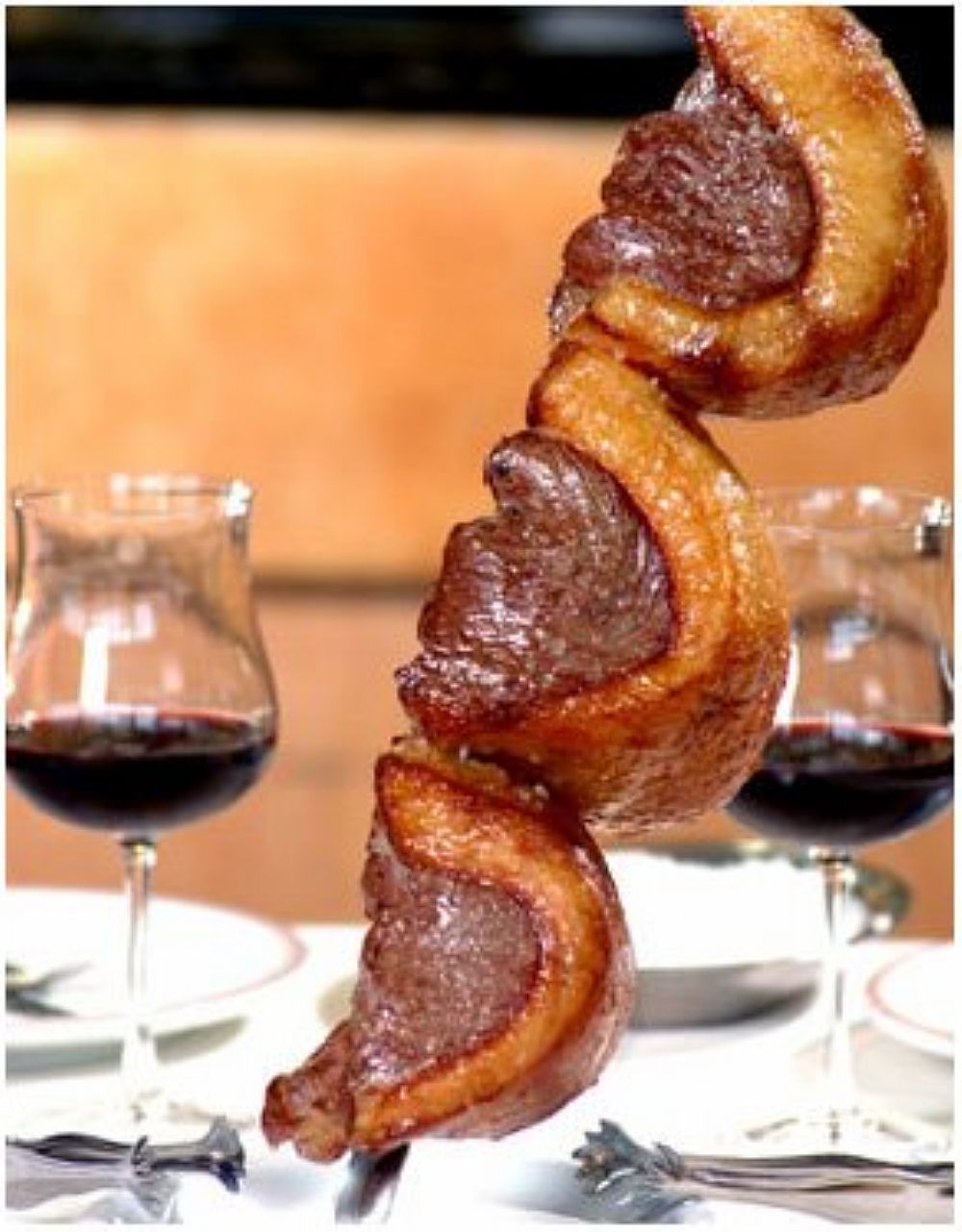 Espeto com 3 pedaços de picanha, apoiado com a ponta sobre a mesa ao lado de duas taças de vinho tinto.