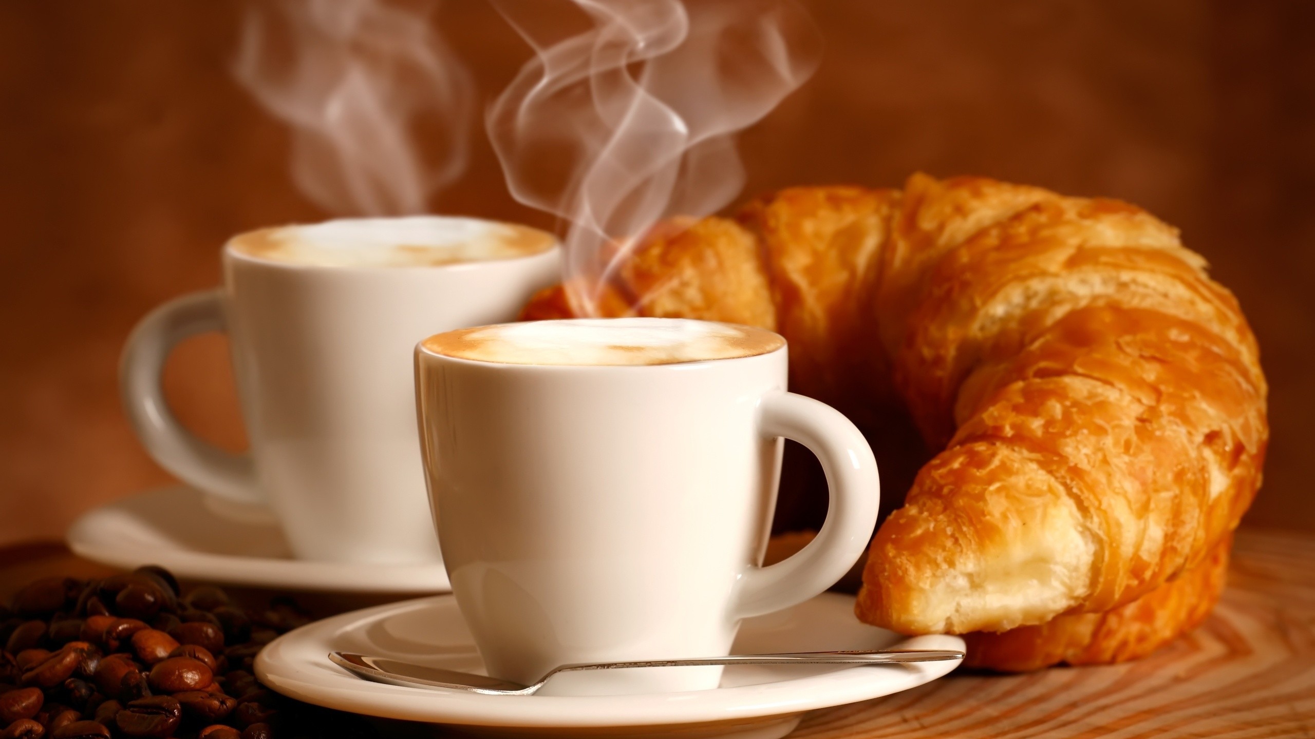 2 Xícaras de café, saindo fumaça ao lado de um croissant, espécie pão folhado e enrolado, servido rechedado ou não.