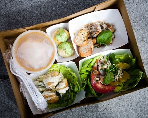 Caixa de papelão, com várias opções de pratos, contidas em potes de papel, com pães, saladas, , pote com sopa, e talheres de plástico