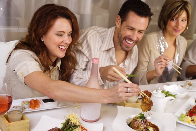 Numa mesa de jantar, um homem vestindo camisa esporte, e de barba, está entre duas senhoras , eles degustando comida japonesa .japonesa - O