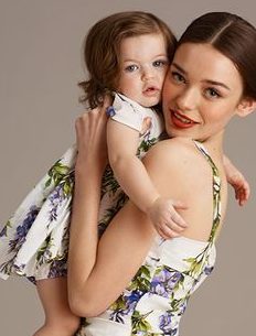 Mulher usa vestido , cor branca, com detalhes em flores roxas e azuis. O modelo usa alças finas. No colo dela, uma pequena menina com o mesmo modelo da mãe.