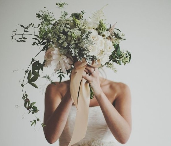 mulher usa vestido de noiva, estilo tomara que caia, ela segura um buquê de flores junto ao seu rosto.