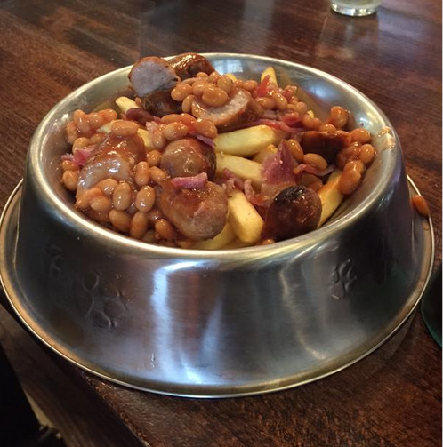 Uma vasilha redonda e funda de alumínio própria para alimentos de cachorros e gatos apresenta uma feijoada com batatas fritas e pedaços de paio e linguiça