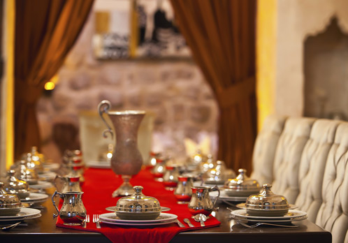 Uma mesa retangular colocada com toalha vermelha e cerca de 8 ou 9 pratos com a uma colhe de prata por cima de cada um deles estão enfileirados simetricamente . Ao fundo uma jarra de prata trabalhada completa o visual.