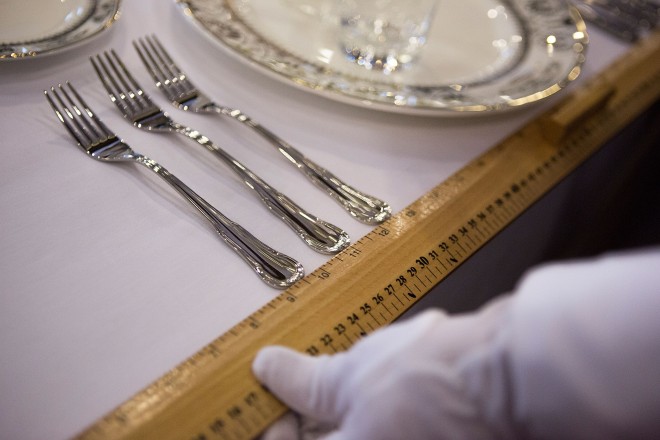 Foto em close de uma mesa colocada com uma mão de luvas brancas segurando uma régua amarela para medir a distancia entre um prato e outro.
