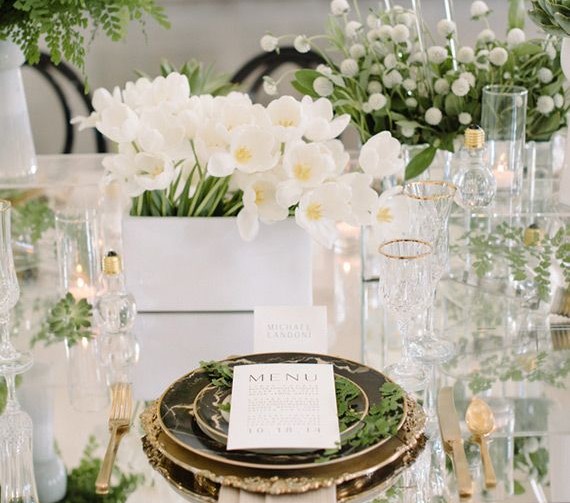 Mesa de jantar, decorada em tons de branco e amarelo, flores, copo de leite, o guardanapo está sob os pratos e se estende para o chão. O supla em tons dourados , e pratos de porcelana , cor preta e frisos dourados.