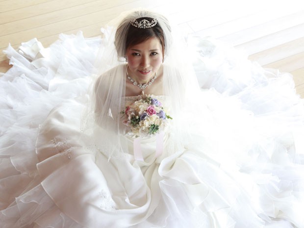Mulher jovem japonesa, está sentada no chão, usando vestido de noiva, bem rodado, usa tiara, e segura um buque de flores junto ao rosto.