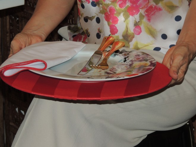 Pessoa sentada, onde não aparece o rosto e em primeiro primeiro plano, está sobre o colo um prato estampado com os talheres que estão apoiados em um apoio de prato vermelho específico para facilitar a refeição sem o auxílio da mesa.