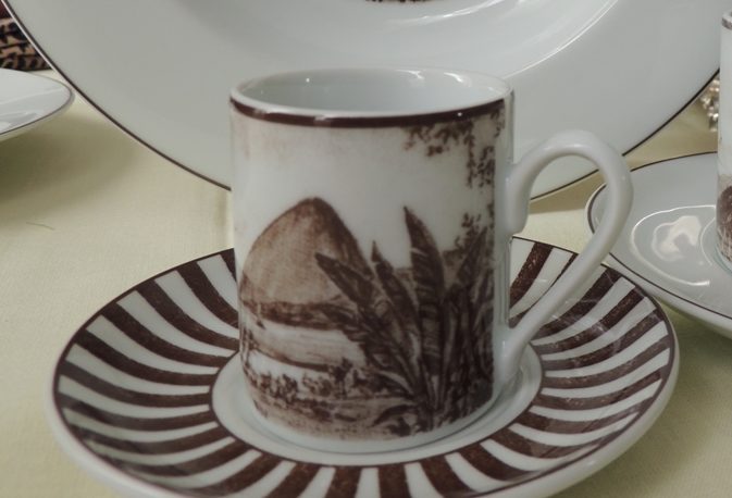 Coleção de porcelana Schmidt, linha Francesca Romana, ao fundo um prato fundo, com figura da Lapa do Rio de Janeiro em sépia, a frente xicaras de cafe e xícara de chá, com imagens o Rio de Janeiro em sépia.