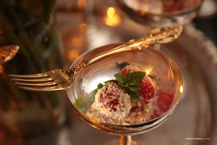 Taça dourada, e um garfo cruzado sobre a borda da taça. Dentro , morangos com uma cobertura de queijo, mel e pimenta.