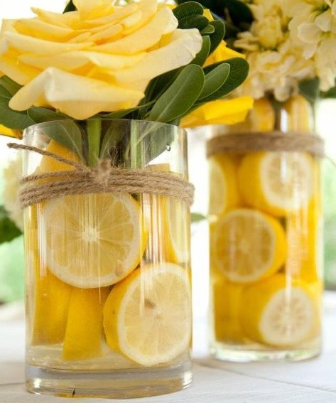 em um jarro de boca larga estão mergulhadas rodelas de limão siciliano amarelas que servem de enfeite junto com a água e compõe a cor do arranjo de rosas amarelas