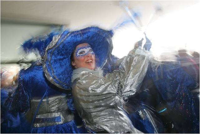 mulher com uma máscara branca pintada na região dos seus olhos e borda azul, usa enorme chapéu azul com bordas prateadas e roupa prateada, canta e brinca num bloco de rua.