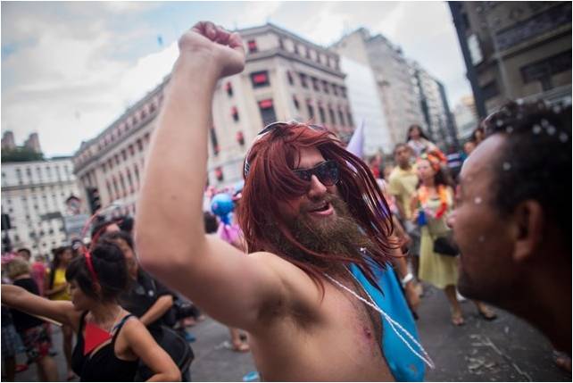 imagem de um homem , folião, usando peruca de cabelos vermelhos usa óculos e está sem camisa, brinca junto aos demais foliões numa avenida.