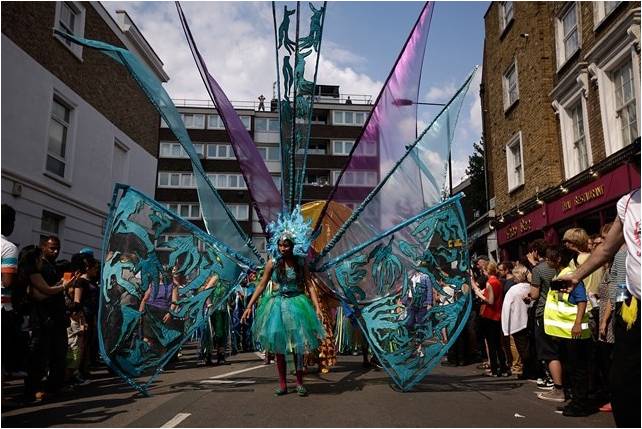 bloco carnavalesco na rua, ao centro uma mulher usando fantasia verde transparente tem em suas costas mastros com bandeiras coloridas. ao redor muitas pessoas apreciam o desfile.