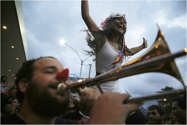 foliões de bloco de rua, um homem com nariz de palhaço toca trombone de vara e uma mulher sorri, com os braços abertos usando top branco com alcinhas, ela usa uma máscara sobre a testa.