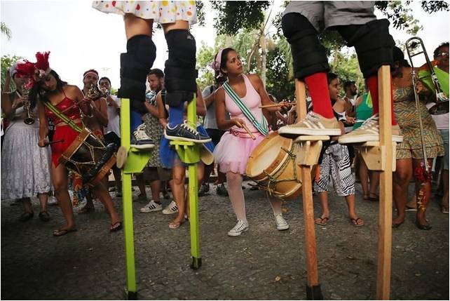 Bloco de rua, pessoas fantasiadas de bailarina sobre pernas de pau. alguns tocam tambores rústicos