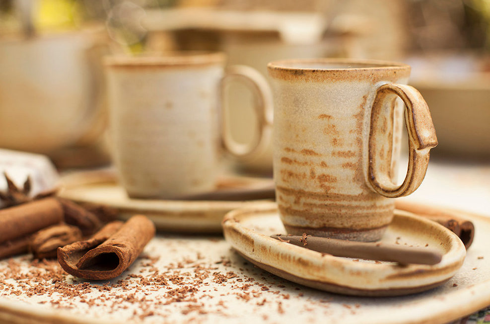 prato em cerâmica com pó de canela e pau de canela ao lado xícaras de cerâmica, alta e na cor de creme com manchas na cor marrom.
